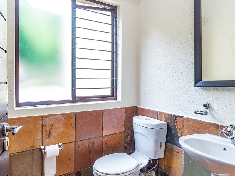 A functional bathroom featuring a pedestal basin, ceramic toilet, and rustic tiled splashback with security bars on the window.