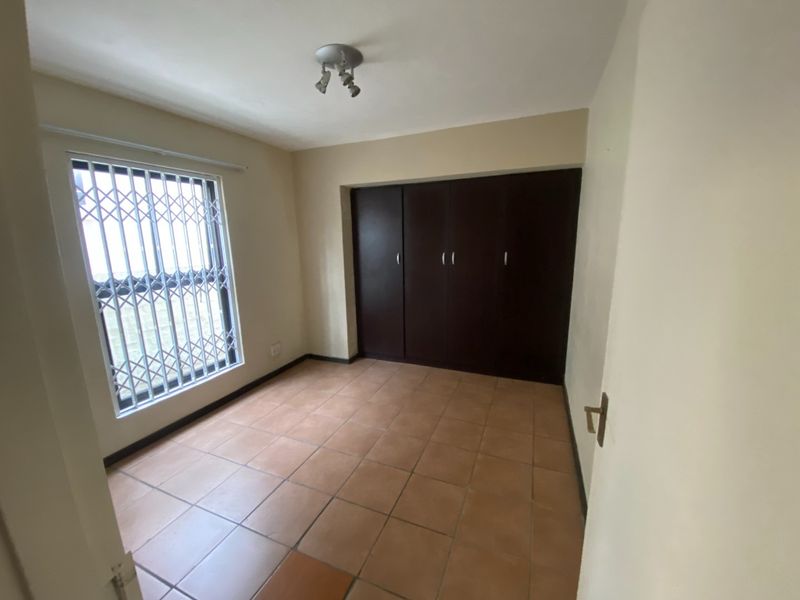 An empty bedroom featuring tiled flooring, built-in dark wood cupboards, and a window fitted with a security gate.