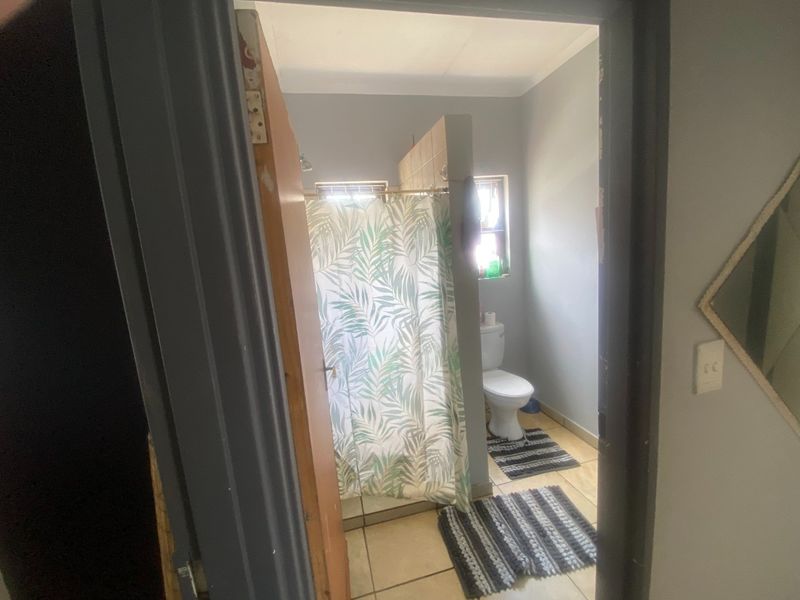 A functional bathroom featuring a shower with a partition wall, a toilet, and tiled flooring, viewed from the adjacent hallway.