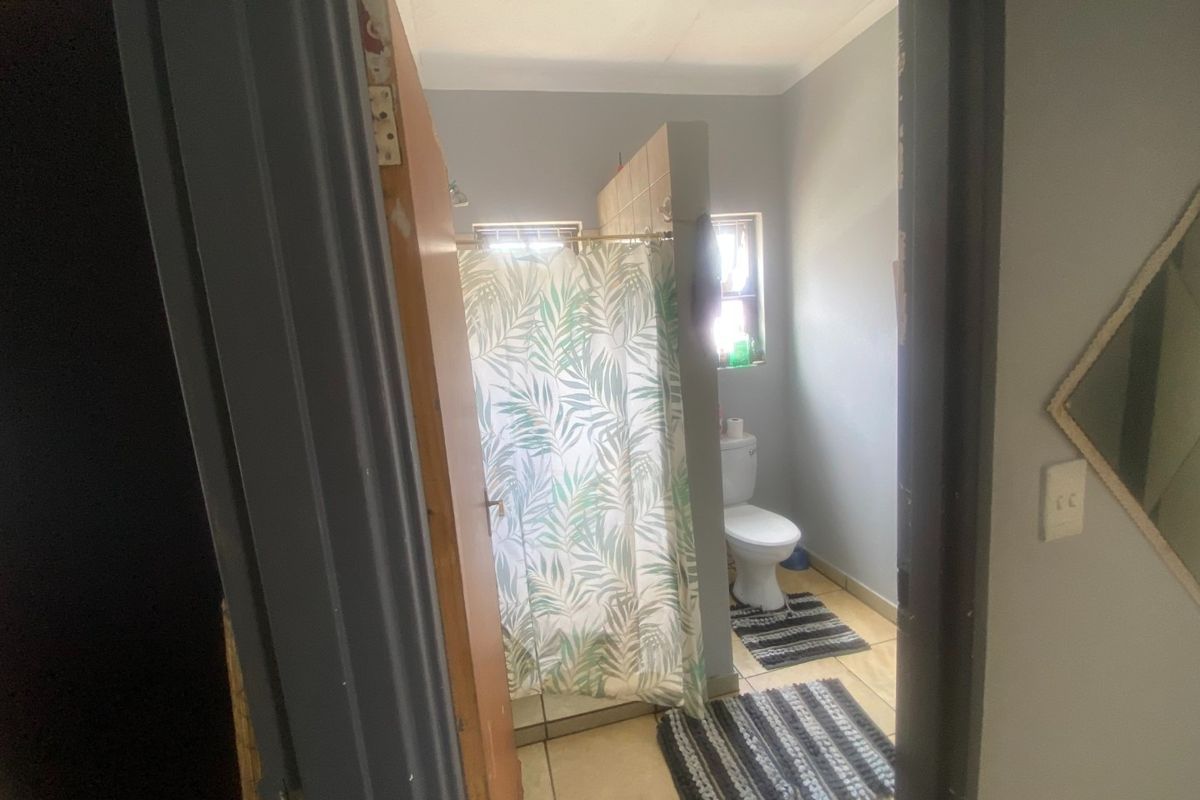 A functional bathroom featuring a shower with a partition wall, a toilet, and tiled flooring, viewed from the adjacent hallway.