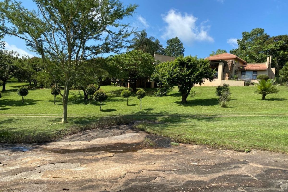 A view of the rear garden featuring a natural rock formation, expansive lawn, mature trees, and a glimpse of the main dwelling with a patio.