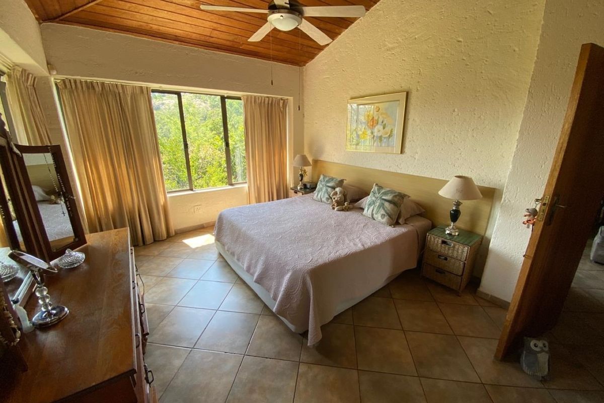 Spacious bedroom featuring a vaulted timber ceiling, ceiling fan, tiled flooring, and a built-in headboard with natural light from large windows.