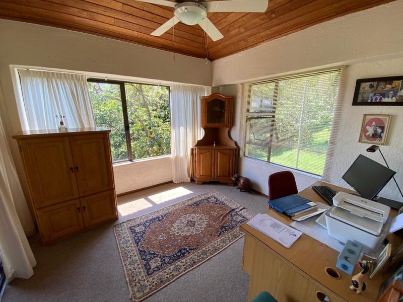 A home office featuring a timber-paneled ceiling, ceiling fan, and large windows providing natural light and garden views.