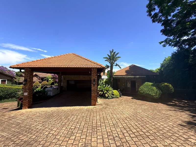 Front exterior view of the property featuring a spacious brick-paved driveway, a covered carport, and well-maintained landscaped garden areas.