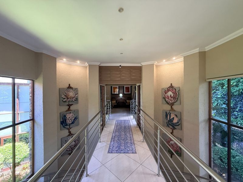 A bright and spacious elevated hallway featuring a modern stainless steel balustrade, tiled flooring, and large windows overlooking the surrounding greenery.