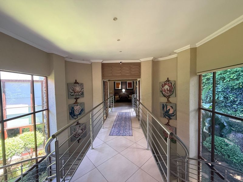 A bright, elevated hallway featuring tiled flooring, stainless steel balustrades, and large windows overlooking the surrounding greenery.