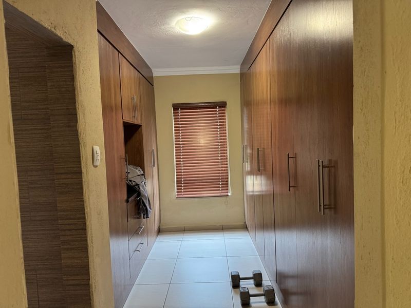 A functional dressing area or bedroom space featuring extensive floor-to-ceiling built-in wooden cabinetry, tiled flooring, and a window with fitted venetian blinds.