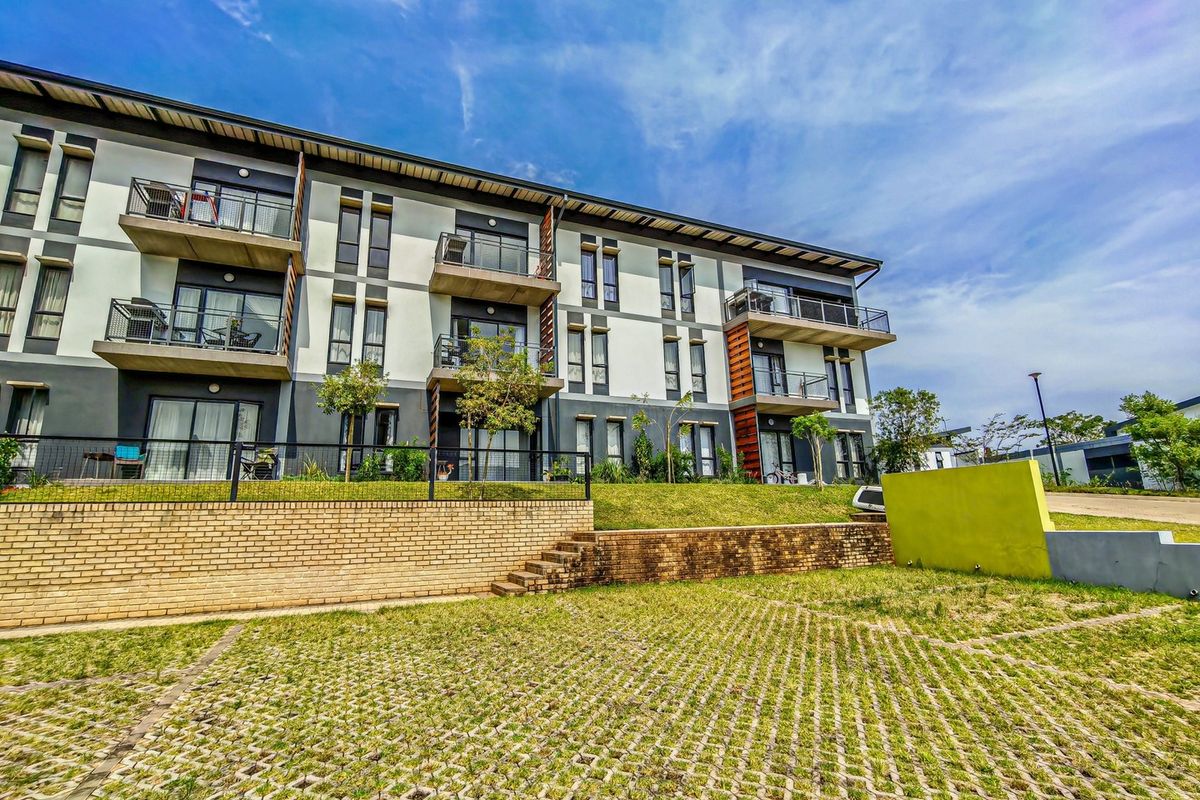 Exterior view of a modern apartment complex featuring private balconies, sliding glass doors, and a landscaped brick retaining wall with paved parking.
