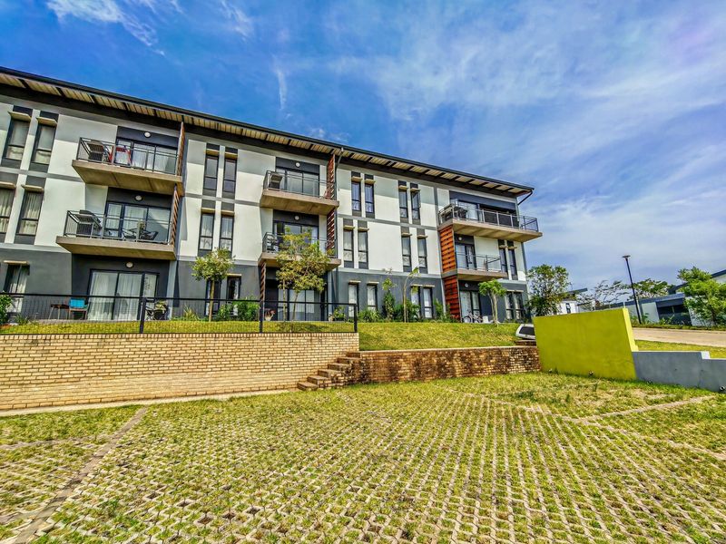 Exterior view of a modern apartment complex featuring private balconies, sliding glass doors, and a landscaped brick retaining wall with paved parking.
