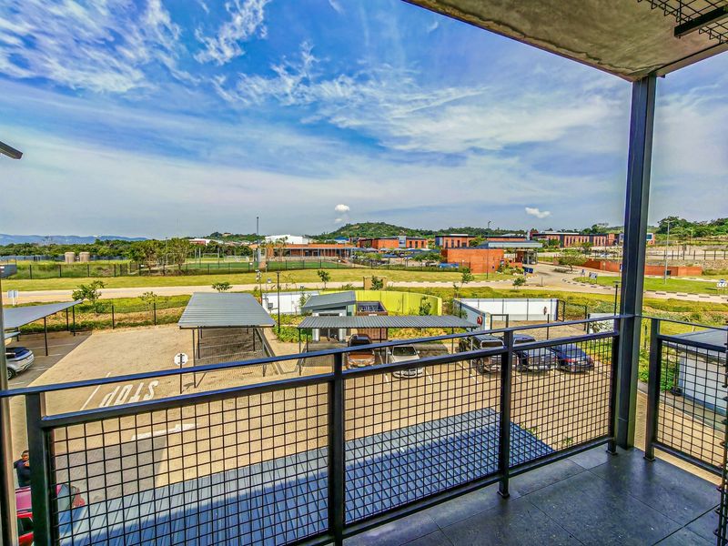 A spacious balcony featuring a metal balustrade and tiled flooring, offering an elevated view over the complex parking area and surrounding landscape.