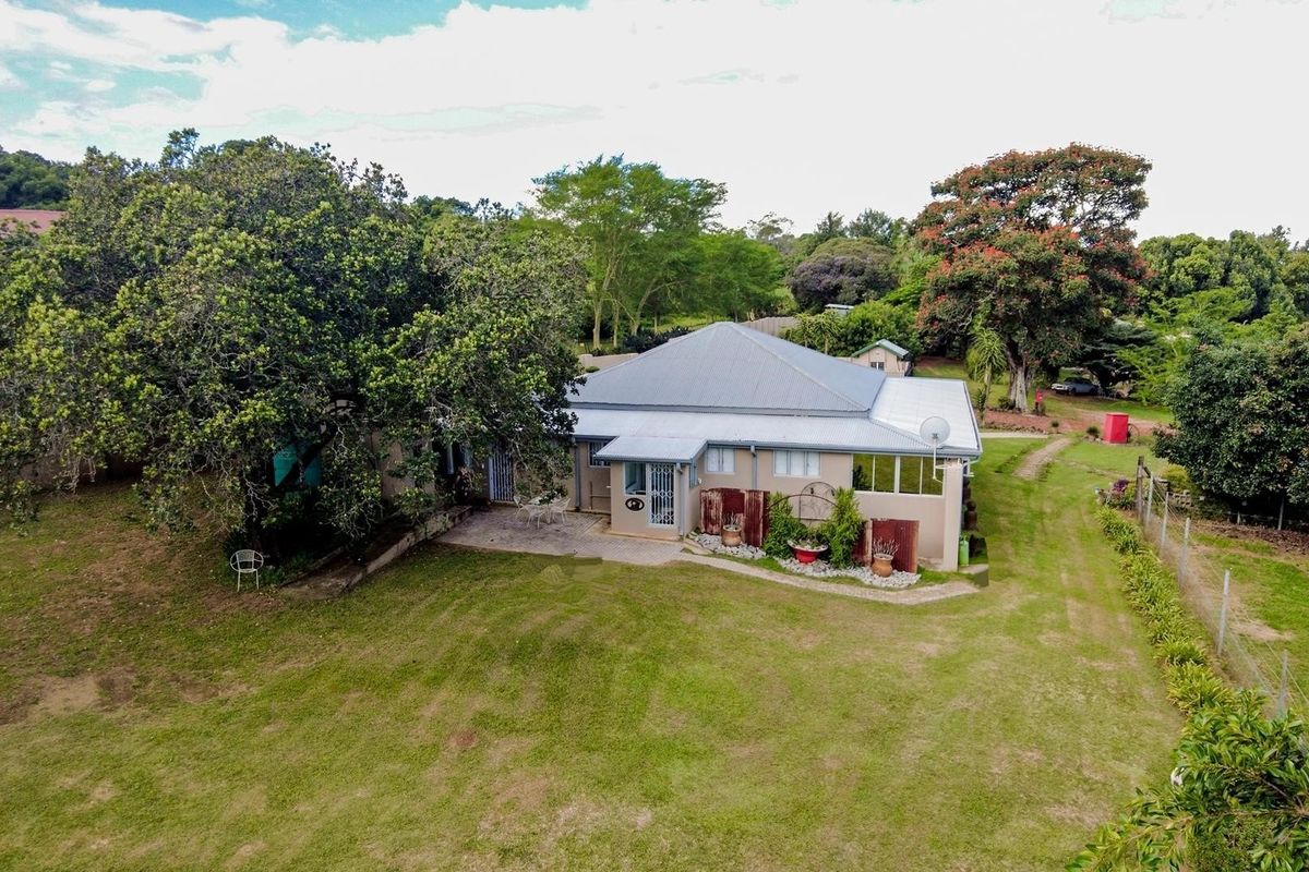 Aerial view of a small holding property featuring a single-story dwelling with a metal roof, paved patio area, and expansive surrounding garden grounds.