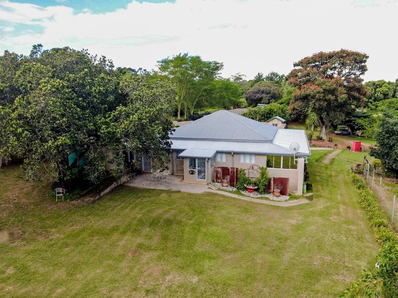 Aerial view of a small holding property featuring a single-story dwelling with a metal roof, paved patio area, and expansive surrounding garden grounds.