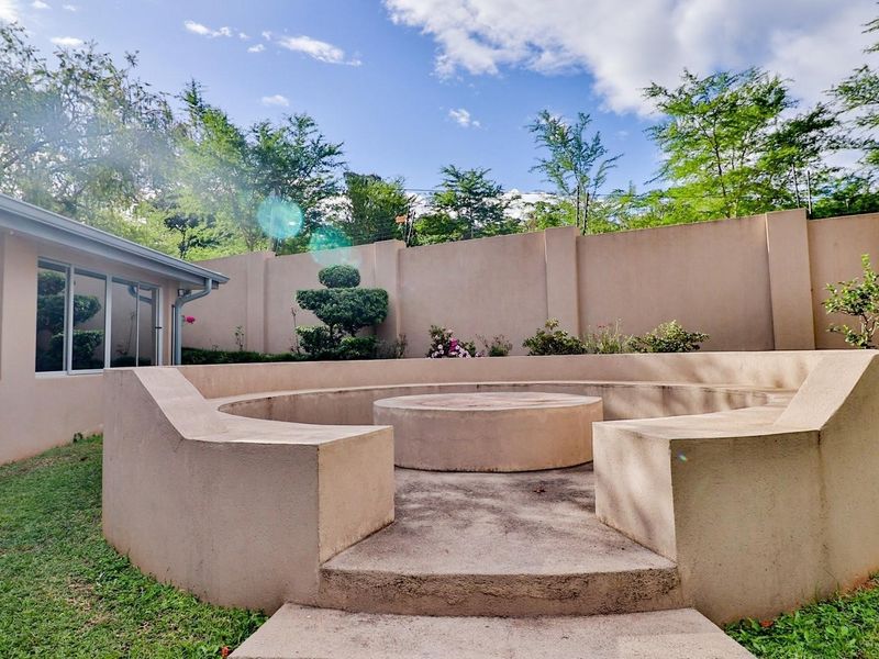 A landscaped garden area featuring a circular built-in concrete fire pit with integrated seating, enclosed by a high perimeter boundary wall.