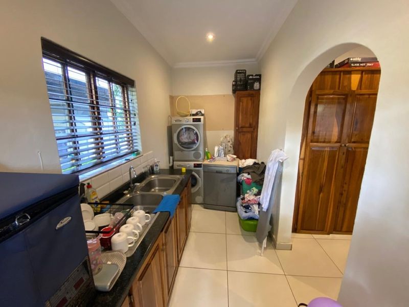 Functional laundry area featuring a double basin sink, tiled splashback, ample wooden cabinetry, and tiled flooring, with an arched entrance to the adjacent space.