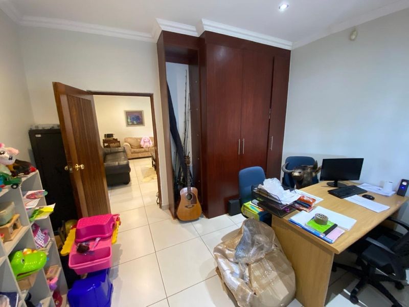 A functional home office space featuring built-in wooden cupboards, tiled flooring, and recessed ceiling lighting, with direct access to the adjacent living area.
