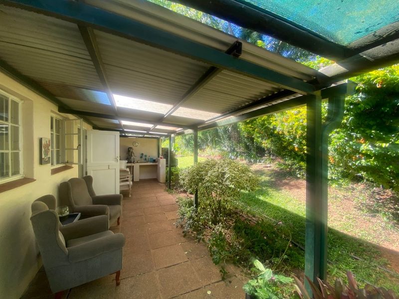 A covered outdoor patio featuring tiled flooring, a built-in counter, and a corrugated roof, overlooking a lush garden area.