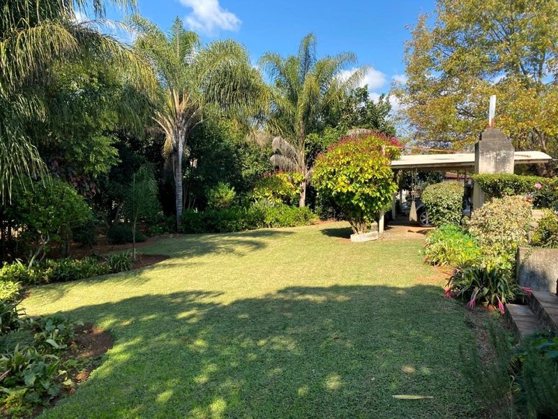 A well-maintained, lush garden area featuring mature trees, manicured lawn, and an adjacent carport structure.