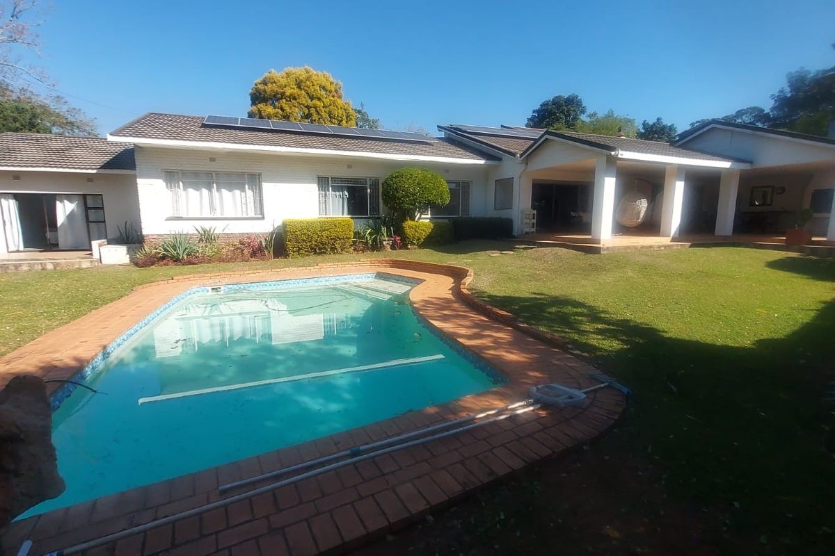 Rear view of the property featuring a swimming pool with brick coping, a covered patio area, and a well-maintained lawn under a tiled roof.