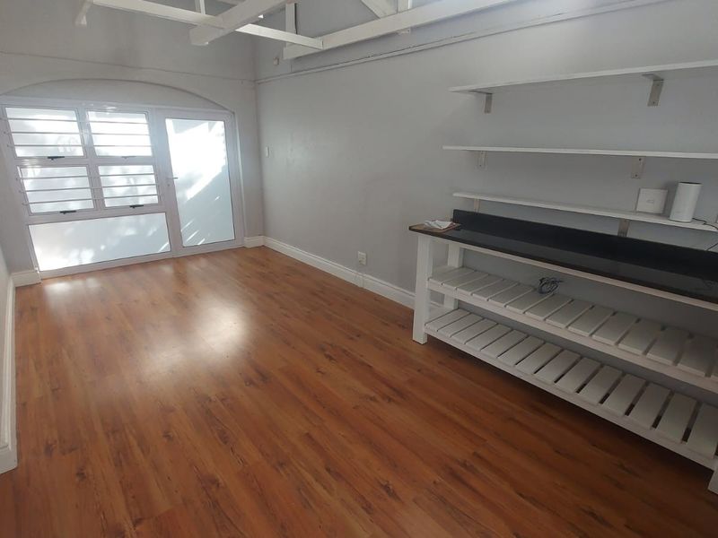 An empty living area featuring laminate flooring, exposed ceiling beams, and built-in shelving with a counter, providing a versatile space with natural light.