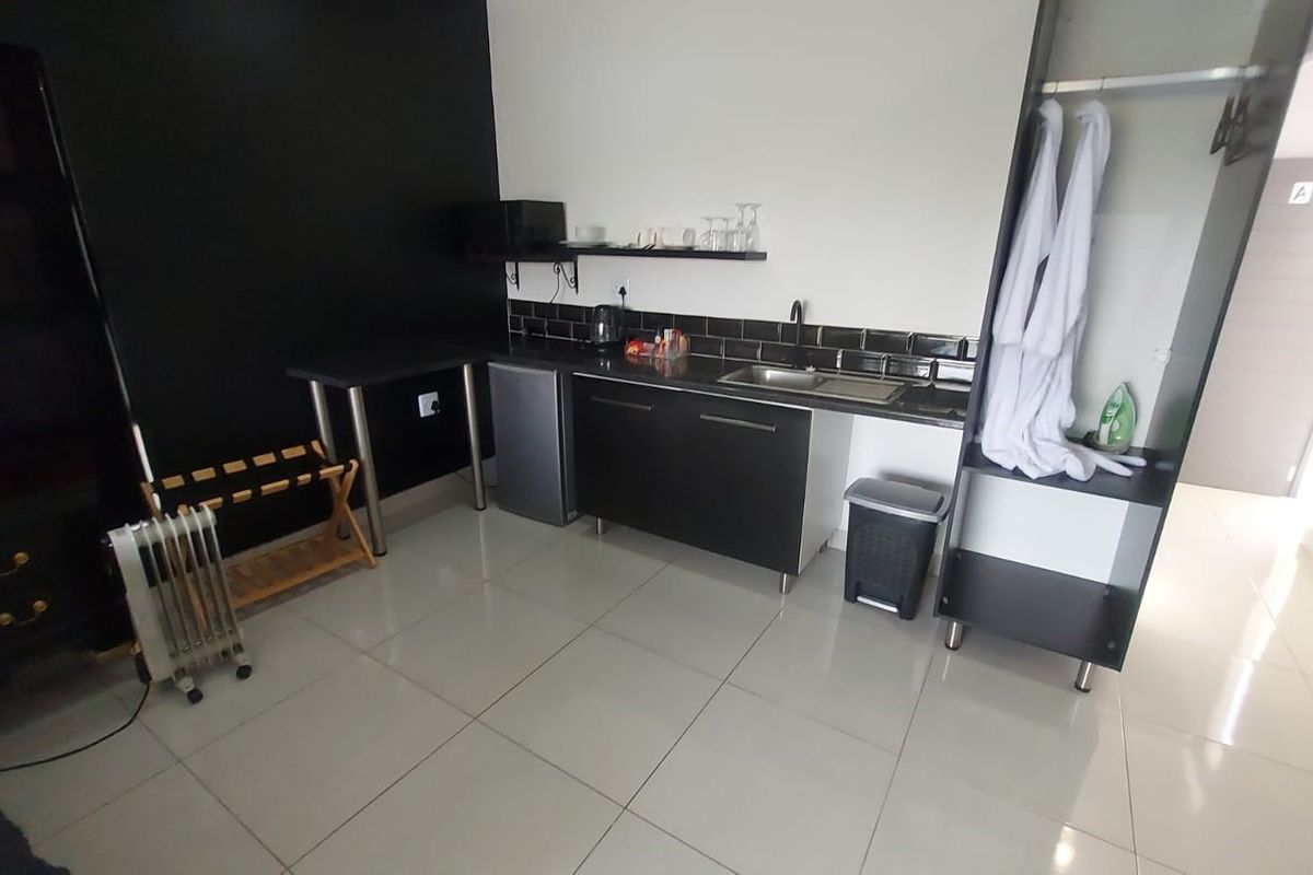 A compact kitchenette area featuring modern dark cabinetry, a sink, tiled backsplash, and an integrated wardrobe unit with tiled flooring.