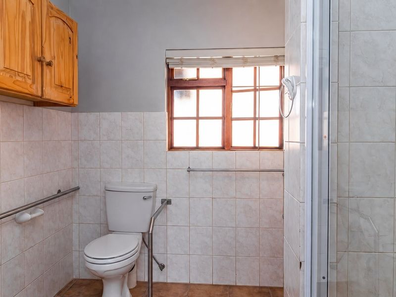 A functional bathroom featuring tiled walls, a shower enclosure, toilet, and wall-mounted wooden cabinetry, equipped with safety grab rails.