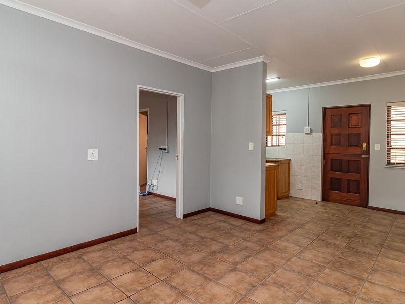 An open-plan living area featuring tiled flooring, neutral wall tones, and a functional kitchen space with wooden cabinetry and direct access to other rooms.