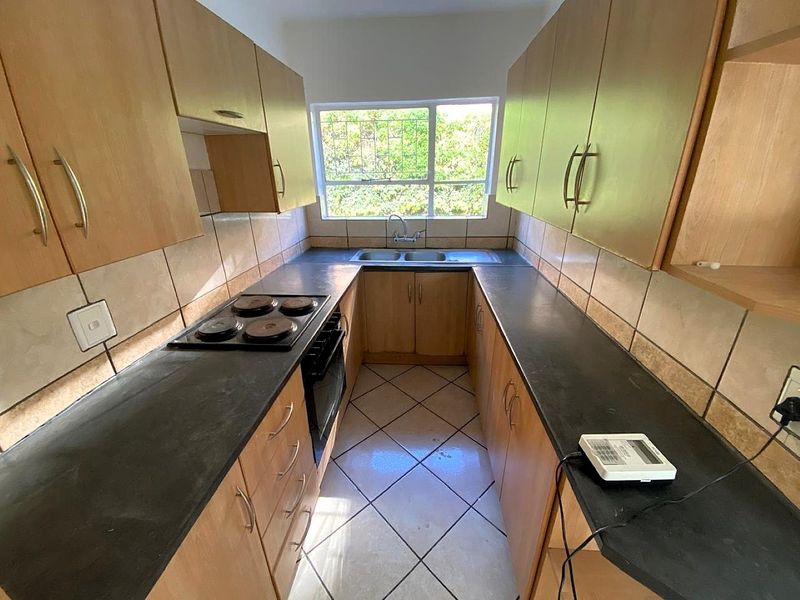 A functional galley-style kitchen featuring light wood-look cabinetry, dark countertops, built-in oven and hob, and a double sink with natural light.