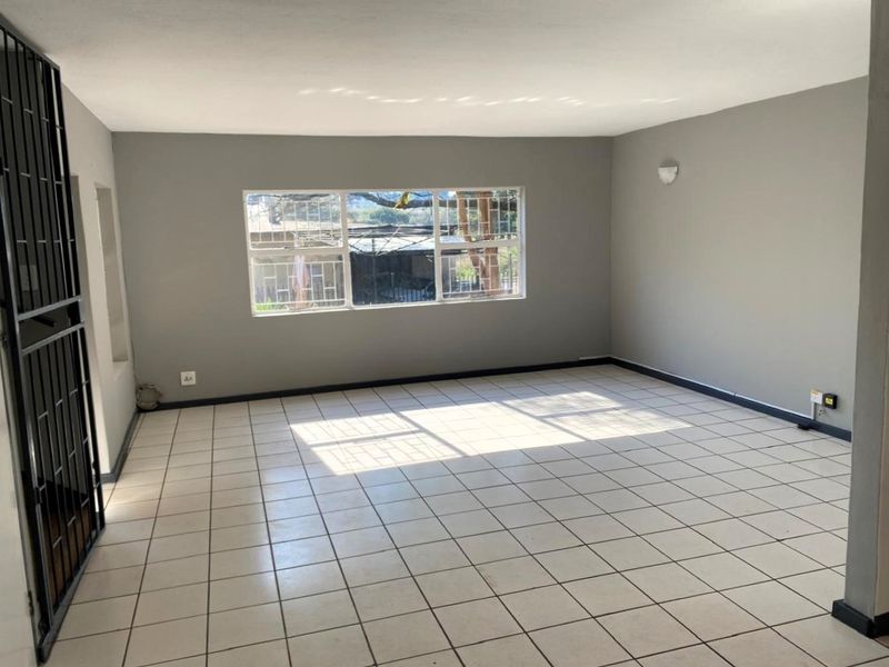 A spacious, empty living area featuring neutral grey walls, white tiled flooring, and a large window with security bars.