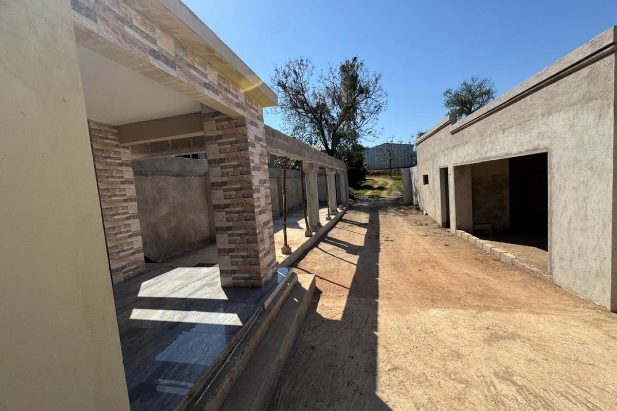 An exterior view of a farm property featuring a tiled patio with stone-clad pillars and unfinished masonry structures along a dirt driveway.