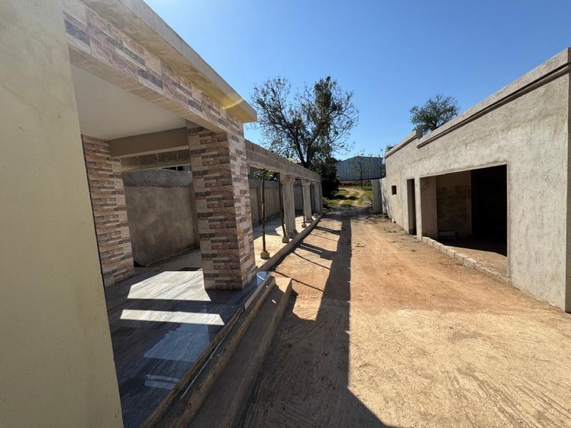 An exterior view of a farm property featuring a tiled patio with stone-clad pillars and unfinished masonry structures along a dirt driveway.