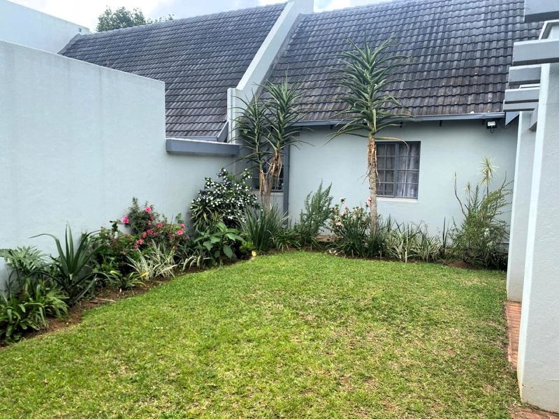 A private rear garden area featuring a small lawn, established garden beds, and a view of the townhouse's rear facade and tiled roof.