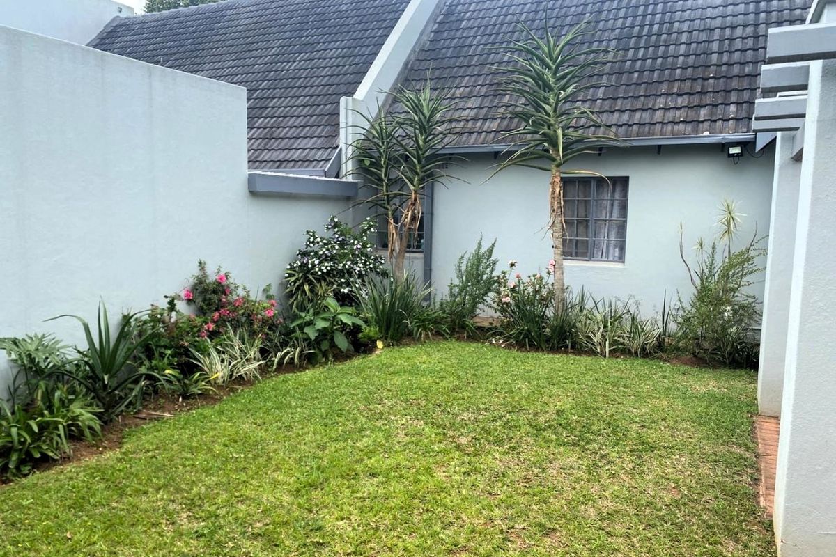 A private rear garden area featuring a small lawn, established garden beds, and a view of the townhouse's rear facade and tiled roof.