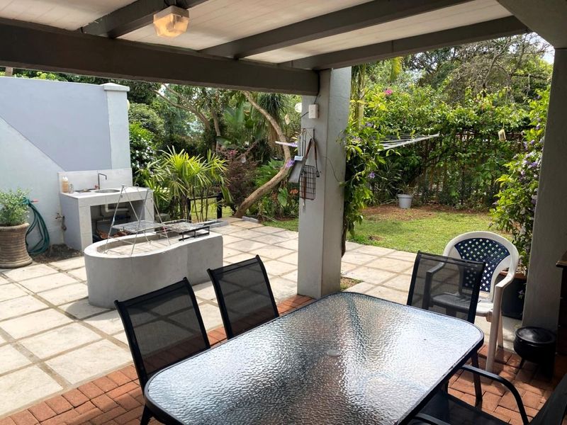 Covered outdoor patio featuring a built-in braai area, sink, and paved flooring, overlooking a lush garden space.
