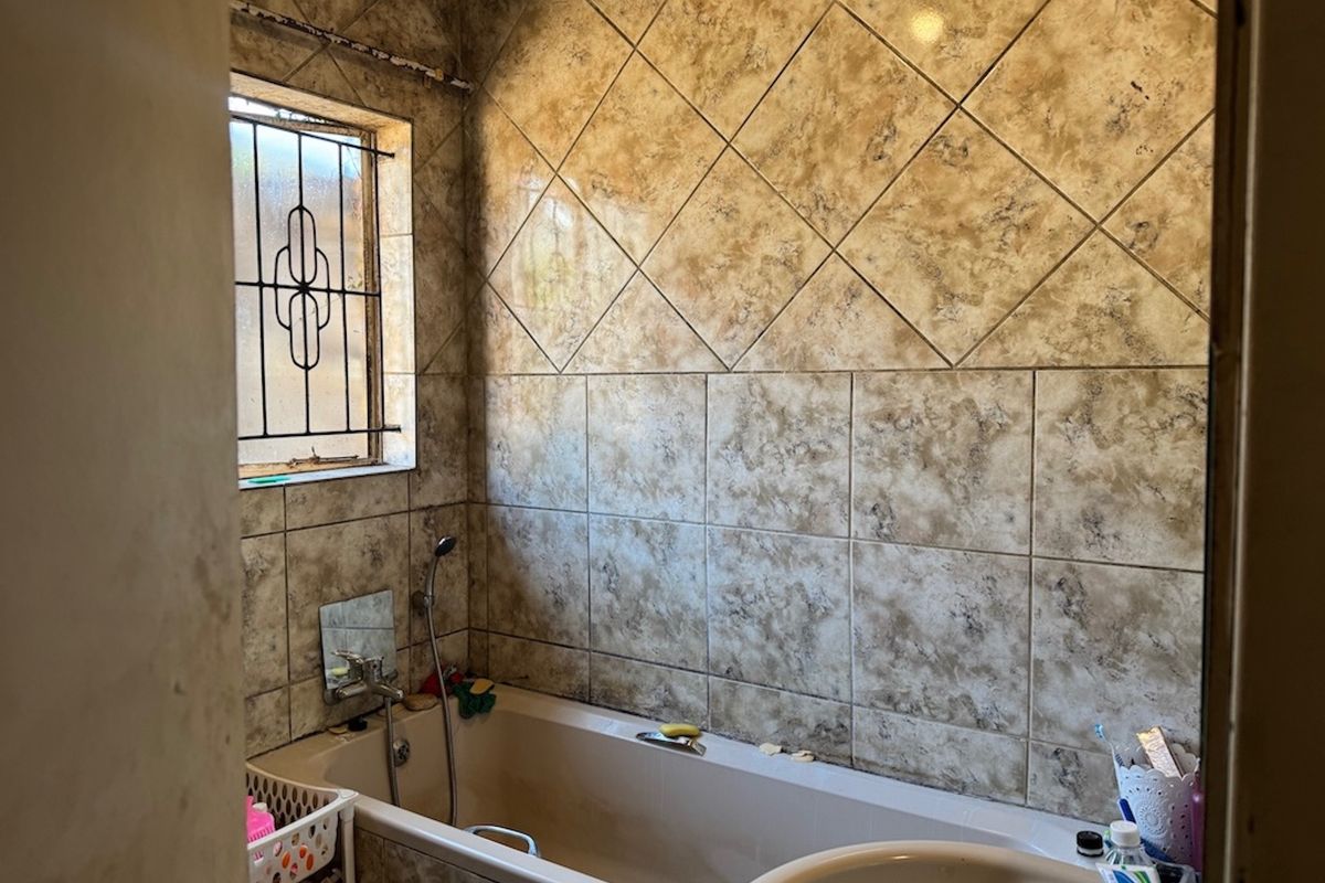 A dated bathroom featuring tiled walls, a bathtub with a shower mixer, and a window with security bars.