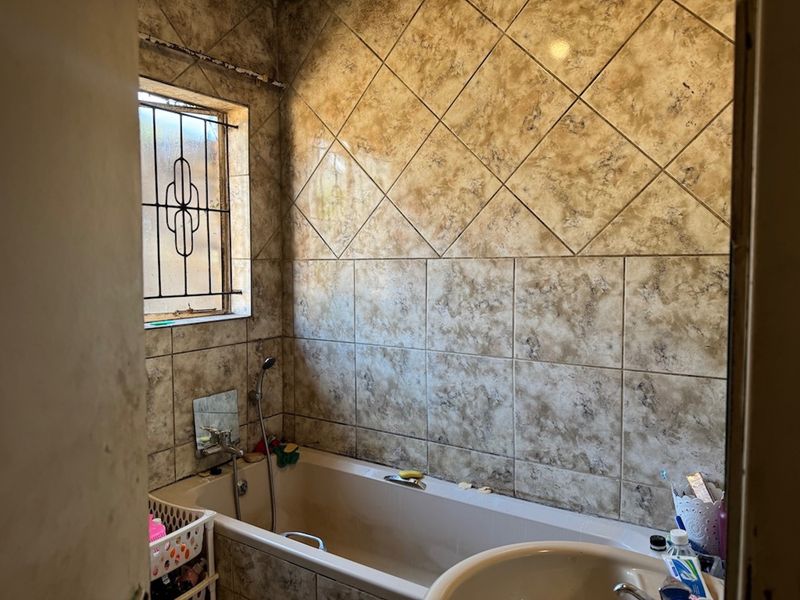 A dated bathroom featuring tiled walls, a bathtub with a shower mixer, and a window with security bars.