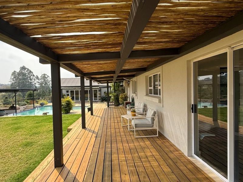 Spacious covered timber deck patio featuring a pergola roof, overlooking a swimming pool and landscaped garden area.