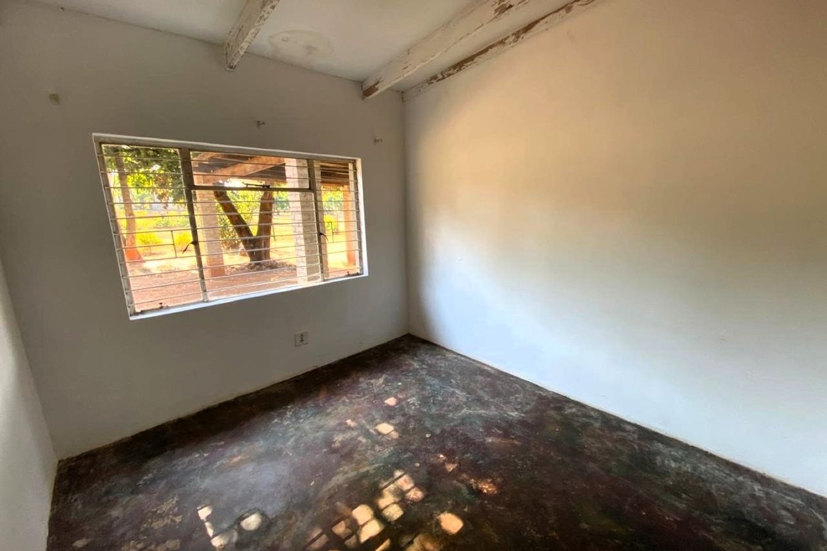 An empty bedroom featuring exposed timber ceiling beams, a steel-framed window with security bars, and unfinished concrete flooring.