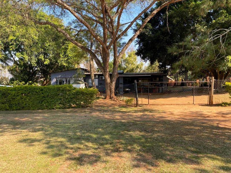 A view of the property frontage featuring a chain-link fence, gated entrance, and mature trees set within a spacious garden area.
