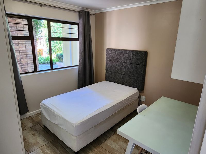 A compact bedroom featuring tiled flooring, a large window with aluminium frames, and a built-in cupboard.