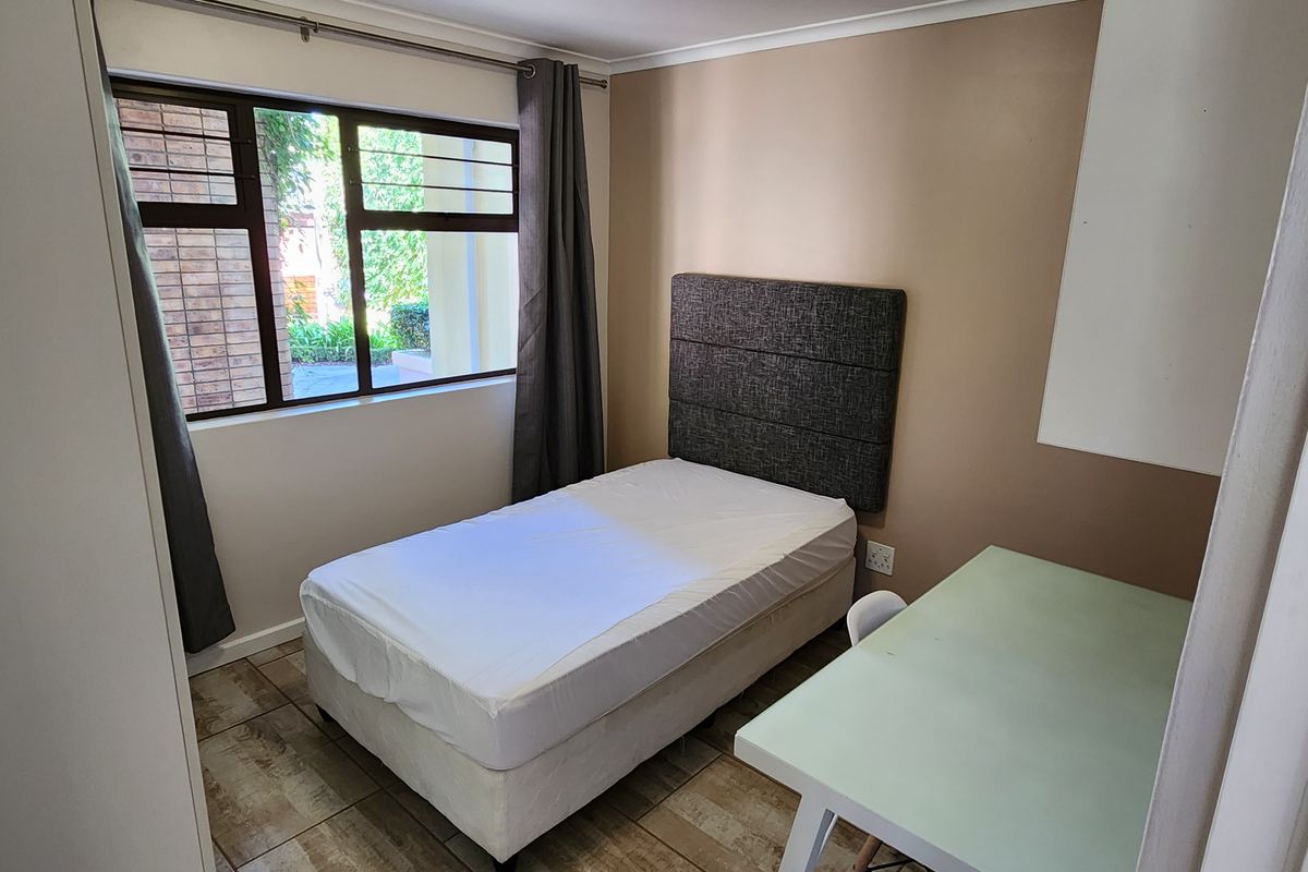 A compact bedroom featuring tiled flooring, a large window with aluminium frames, and a built-in cupboard.