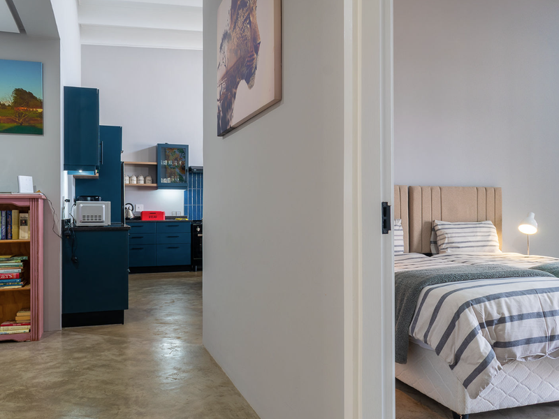 Open plan living area featuring polished concrete floors and a modern kitchen with dark blue cabinetry, seamlessly connecting to an adjacent bedroom.