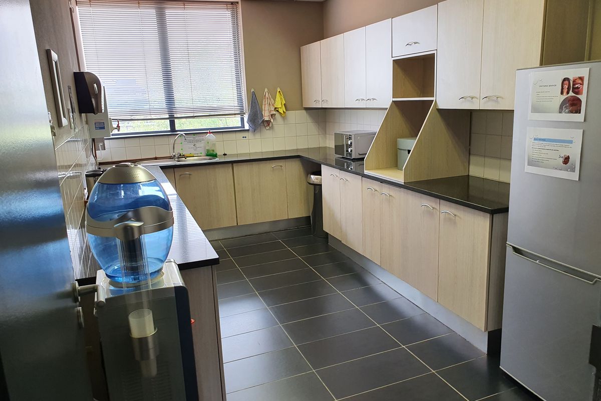 A functional office kitchenette featuring light wood-finish cabinetry, dark countertops, tiled backsplash, and dark floor tiles.