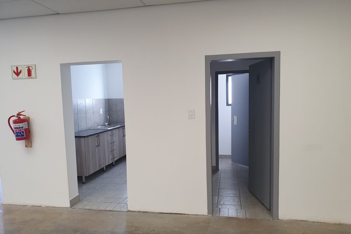 Industrial utility space featuring a kitchenette area with cabinetry and sink, adjacent to a separate restroom entrance, finished with tiled flooring and suspended ceilings.