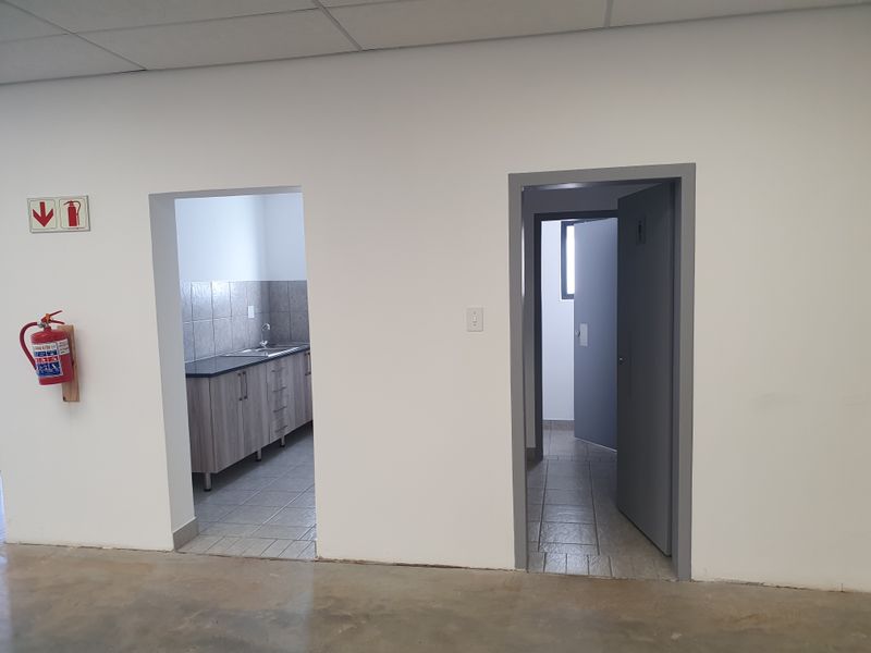 Industrial utility space featuring a kitchenette area with cabinetry and sink, adjacent to a separate restroom entrance, finished with tiled flooring and suspended ceilings.