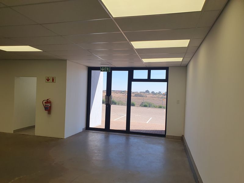 A clean, open-plan industrial entrance area featuring a suspended ceiling, recessed lighting, and a glass door leading to an exterior parking area.