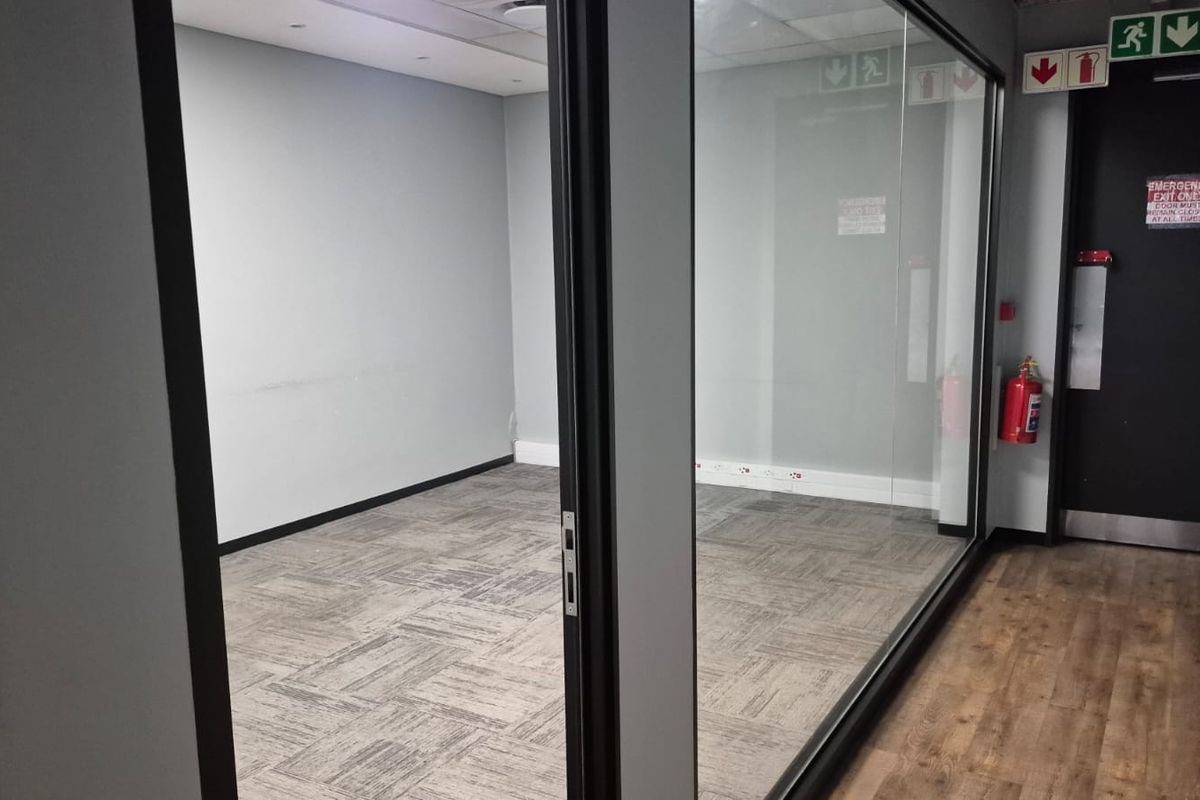 A vacant office space featuring glass partitioning, carpet tiles, and a dedicated emergency exit door with fire safety equipment.