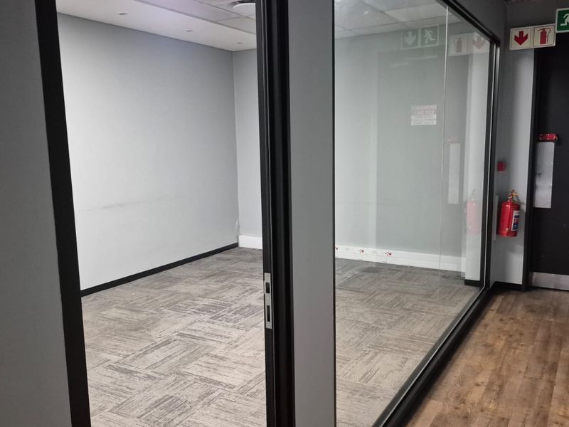 A vacant office space featuring glass partitioning, carpet tiles, and a dedicated emergency exit door with fire safety equipment.