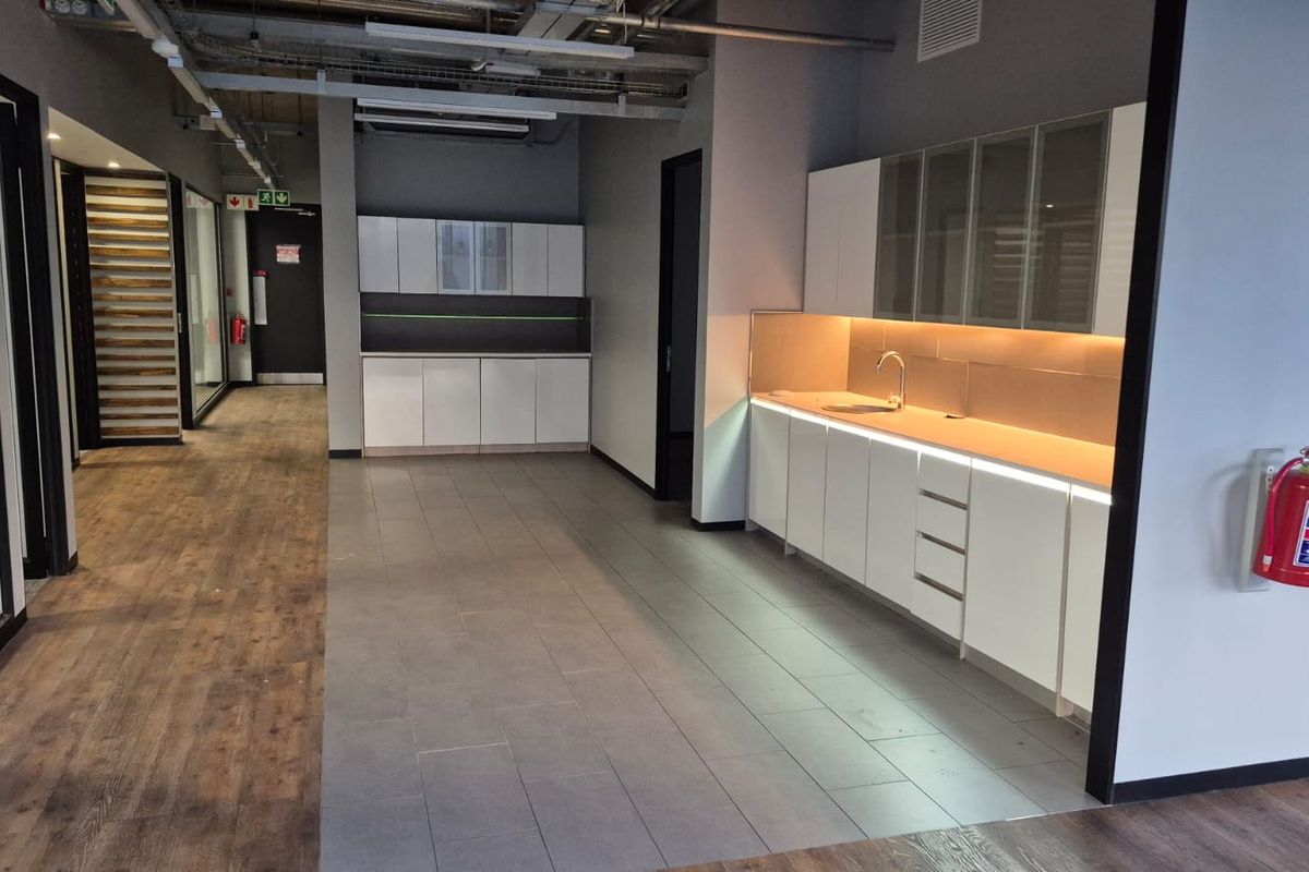Modern office kitchenette featuring sleek white cabinetry, integrated under-cabinet lighting, stainless steel sink, and a mix of tiled and laminate flooring.