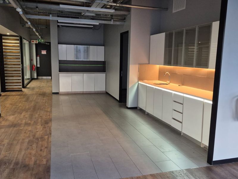 Modern office kitchenette featuring sleek white cabinetry, integrated under-cabinet lighting, stainless steel sink, and a mix of tiled and laminate flooring.