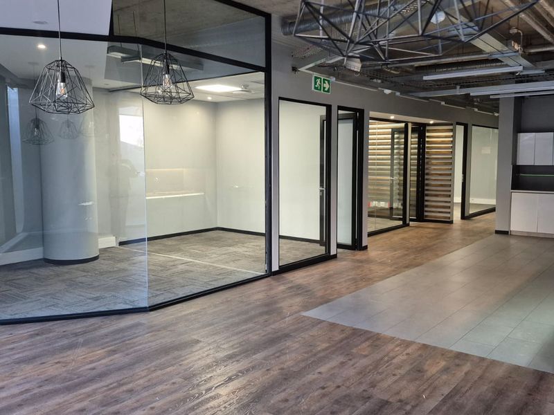Modern open-plan office space featuring glass-partitioned meeting rooms, exposed industrial ceilings, and a compact built-in kitchenette area.